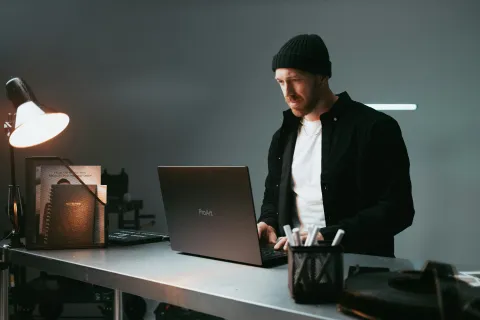 Man wearing beanie and black jacket doing cybercrime on a laptop at a metal desk under a desk lamp.