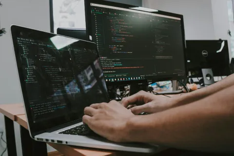 Person coding on a laptop with multiple monitors displaying programming code in a modern office environment.