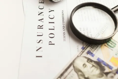 Magnifying glass on a cyber insurance policy document with US hundred-dollar bills partially visible beneath it.