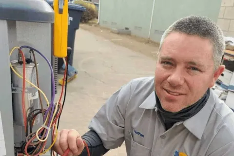Technician in uniform testing HVAC system wiring outdoors with multimeter, smiling at camera