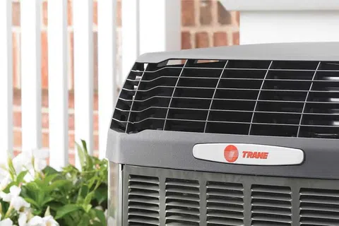 Close-up of a Trane outdoor HVAC unit with a brick wall and plants in the background.