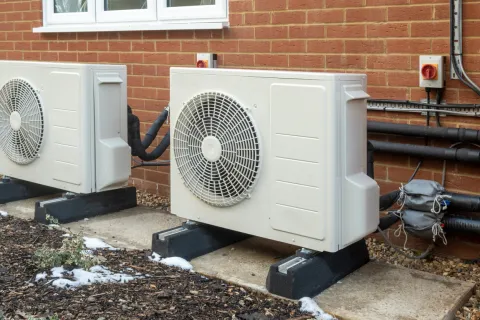 Two large outdoor air conditioning units installed on concrete slabs next to a brick wall with piping and wiring.