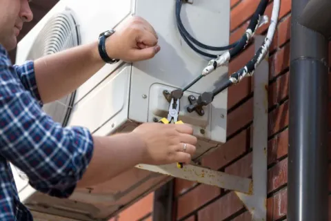 Technician repairing or installing an outdoor air conditioning unit on a brick wall using pliers.
