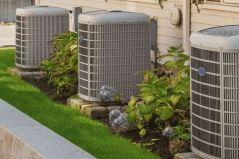 Three outdoor air conditioning units beside a house with green plants and flowers in a landscaped garden bed.