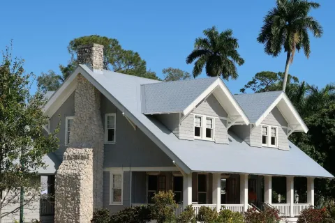 Gray craftsman-style house with stone chimney and large porch surrounded by greenery and palm trees