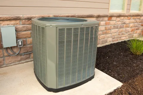 Outdoor central air conditioning unit installed on concrete slab next to brick house wall and garden bed.