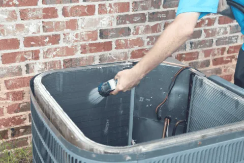 Technician cleaning HVAC condenser coils with a brush spray against a brick wall background outdoors.