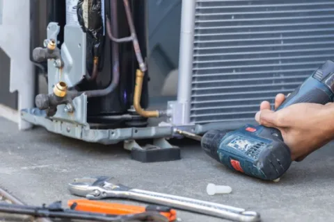 Technician using a cordless drill to repair an HVAC unit on the ground with tools nearby in focus.