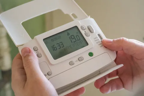 Hands adjusting a white digital thermostat displaying temperature and time settings on a clear screen.