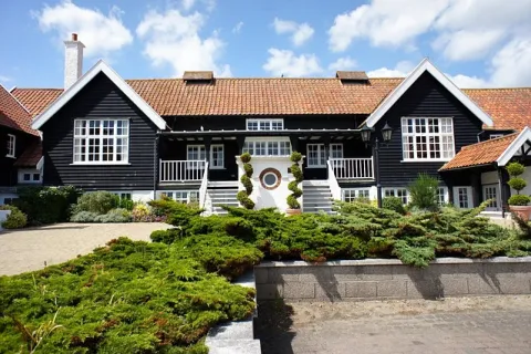 Black and white house with tiled roof surrounded by greenery under a partly cloudy blue sky.