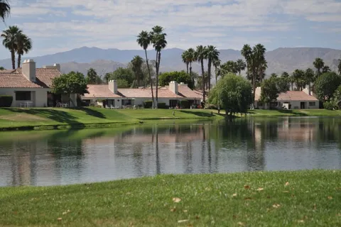 Lakeside residential community with palm trees, green lawns, and mountains in the background under a partly cloudy sky