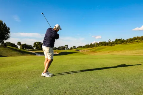 Golfer in navy shirt and white shorts swinging club on green golf course under clear blue sky