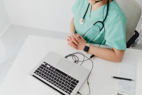 Healthcare professional wearing scrubs with stethoscope and headset using laptop for telemedicine consultation.