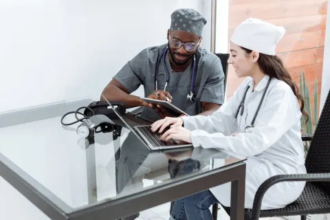 Two healthcare professionals working on a laptop and tablet together at a glass desk in a medical office.