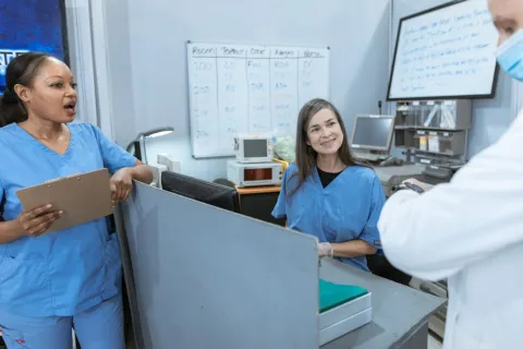 Healthcare workers in blue scrubs discuss patient care at a hospital nurse station with medical equipment visible.