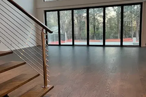 Modern interior with wooden flooring, metal railing staircase, and large floor-to-ceiling windows showing a forest view.