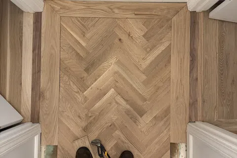 Person standing on newly installed herringbone wood floor with tools laid out, door frames visible on sides.