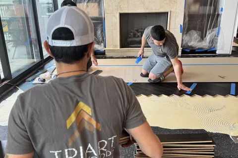 Two workers installing dark hardwood flooring in a modern living room with a fireplace and large windows.