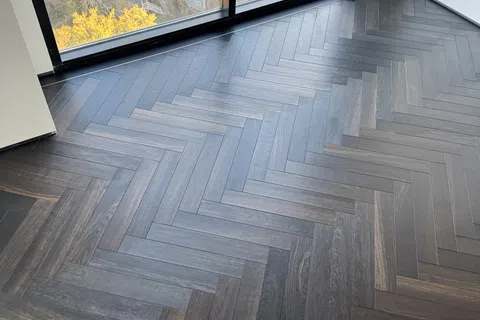 Dark wood herringbone patterned floor in a modern room with large floor-to-ceiling window overlooking autumn trees.