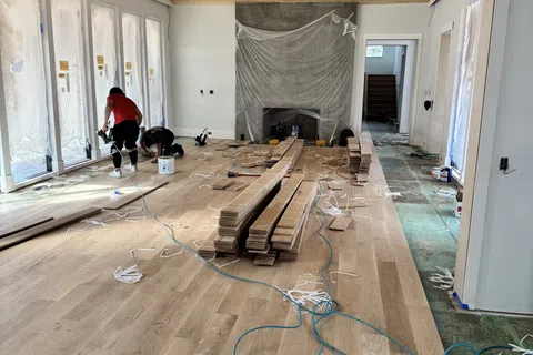 Workers installing wooden flooring in a spacious room with vaulted wood ceiling and covered fireplace.
