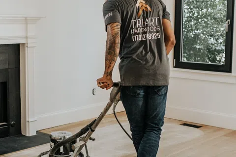 Worker using floor sander to refinish hardwood floors in a bright living room with large windows.