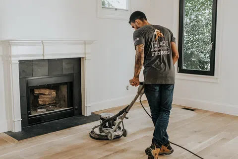 Man sanding hardwood floor near fireplace in bright room with large window and white walls