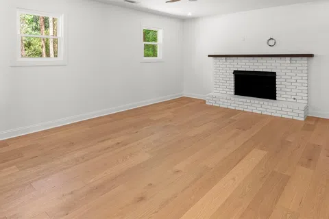 Bright empty room with hardwood floors, white brick fireplace, two windows, and ceiling fan.