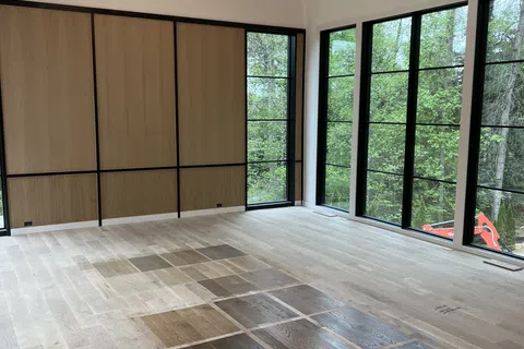 Modern bright room with large black-framed windows, wood floor samples, and an elegant chandelier under arched ceiling.