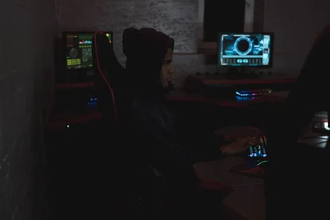 Person in dark hoodie working on computer with futuristic digital interfaces in dimly lit room.