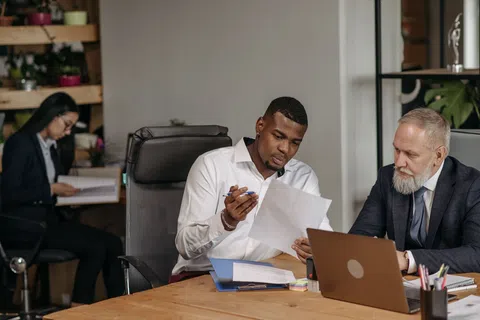 Two businessmen reviewing documents at a desk with laptop while a woman works in the background office