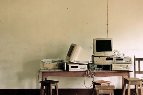 Old computer monitors and desktops stacked on a wooden table with stools and a chair in a sparse room