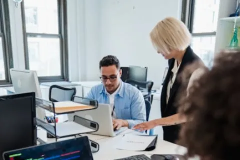 Software developers collaborating in a bright office, with code on a laptop and discussion over documents.