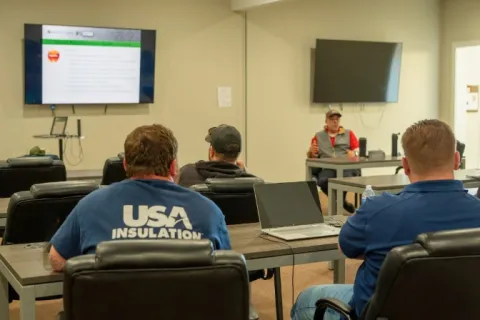 Workers attend a training session in a conference room with presentation slides and laptops visible.