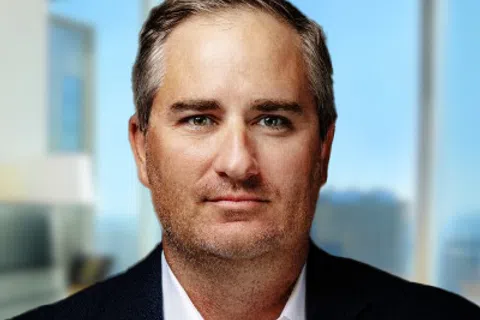 Confident middle-aged man with gray hair wearing a black blazer and white shirt in a modern office setting.