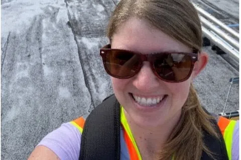 Smiling woman in safety vest and sunglasses takes selfie on industrial rooftop with pipes and equipment in background