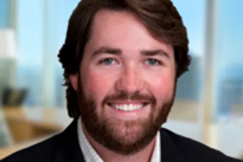 Smiling man with brown hair and beard wearing a suit in a modern office with large windows and blue sky outside