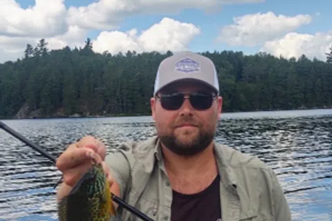 Man wearing sunglasses and a cap holding a fishing rod with a caught fish on a lake with trees and clouds in the background