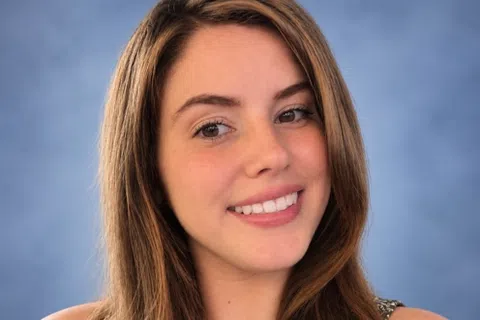 Young woman with long brown hair smiling against a soft blue background wearing a patterned sleeveless top