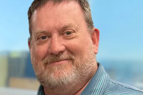 Middle-aged man with short hair and beard wearing a blue striped polo shirt smiling against a blurred outdoor background.