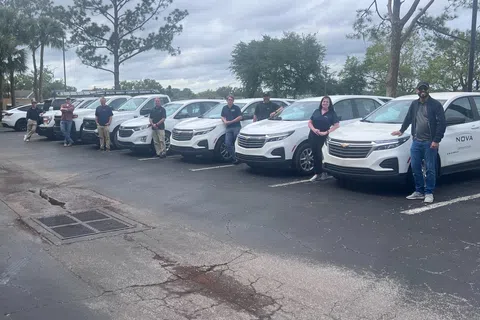 Team of eight people standing beside a row of eight identical white SUVs in a parking lot under cloudy sky.