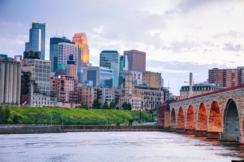 Minneapolis skyline with Stone Arch Bridge over the Mississippi River at sunset under cloudy sky.