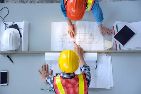 Construction workers in helmets and vests reviewing architectural blueprints on a table with tools and devices