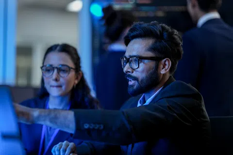 Two professionals in glasses collaborating on a digital project with a computer screen in a dark office.