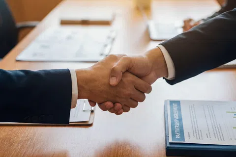 Two business professionals shaking hands over a wooden table with partnership agreement documents visible