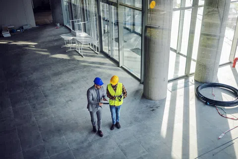 Two construction professionals in hard hats reviewing plans inside a large, modern unfinished building.