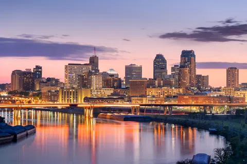 City skyline at twilight with illuminated buildings and a bridge over calm river reflecting lights. Clear sky with soft clouds.