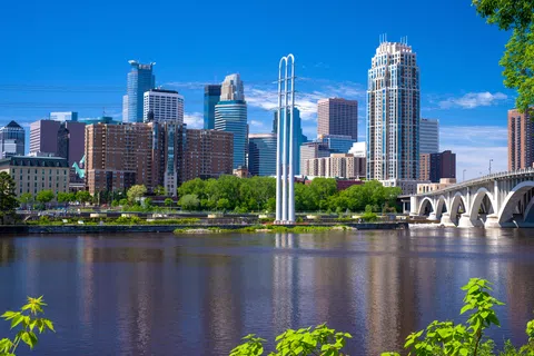 Minneapolis skyline with Stone Arch Bridge and Mississippi River under a clear blue sky with green trees.