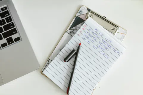 Clipboard with handwritten to-do list and pen next to laptop on a white desk workspace.