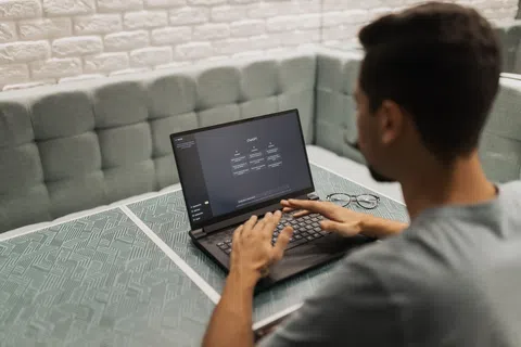 Man typing on laptop with checklist on screen in modern indoor cafe setting with cushioned bench.