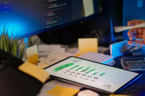 Digital tablet displaying a green highlighted to-do list on a desk with sticky notes, keyboard, and calculator.